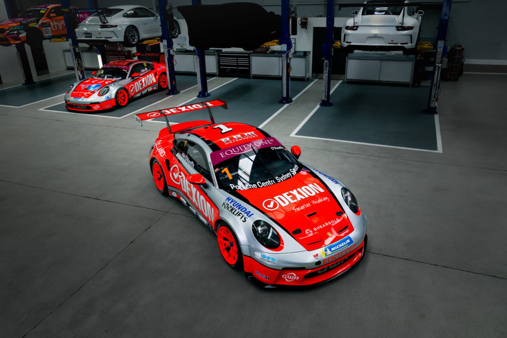 A bright red and silver Porsche race car with Dexion and GutsKorn branding sits in the foreground of a clean automotive workshop. Behind it, several white Porsche sports cars are elevated on hydraulic lifts, and another red race car is parked to the left. The scene is well-lit, highlighting the racing livery, large rear wing, and the organised garage environment.