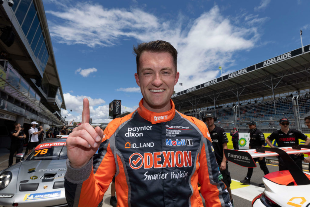 Dexion Victoria sponsored Porsche Carrera Cup driver Dylan celebrating on the Adelaide circuit grid after a race, wearing a Dexion-branded racing suit and raising one finger to signal first place.