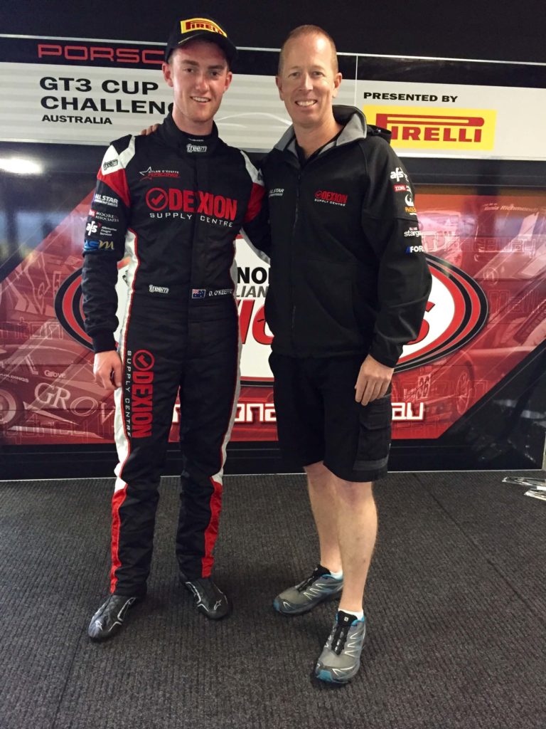 Young racing driver Dylan in a Dexion-branded Porsche GT3 Cup suit standing with team representative at a Porsche GT3 Cup Challenge Australia event.